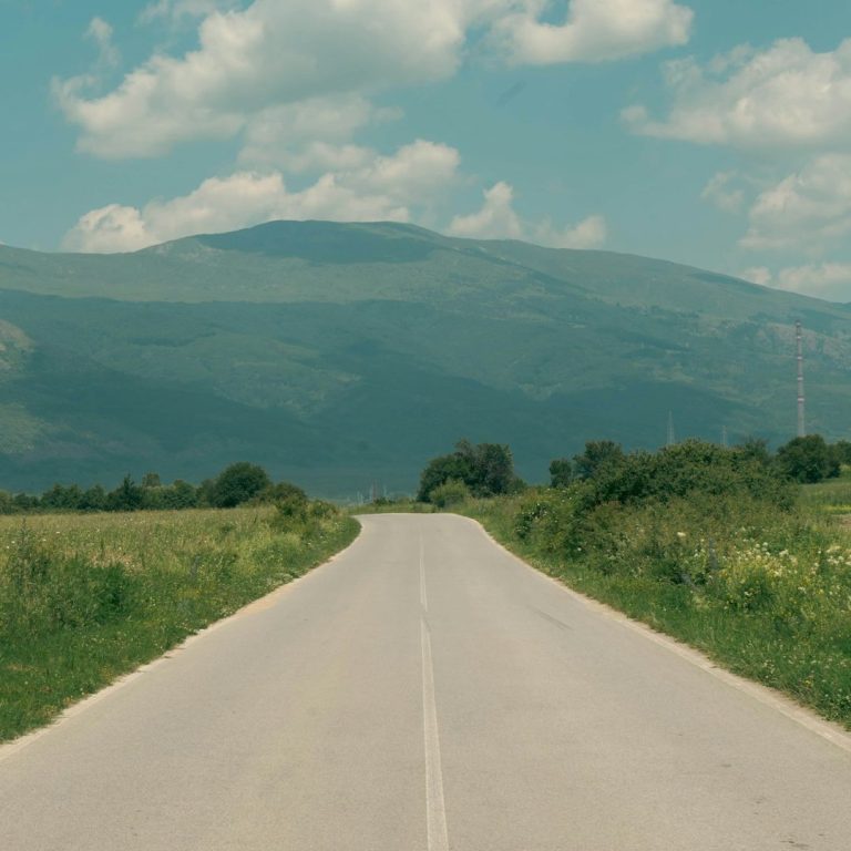 Eine gerade Straße führt durch eine grüne Landschaft mit Bergen im Hintergrund.